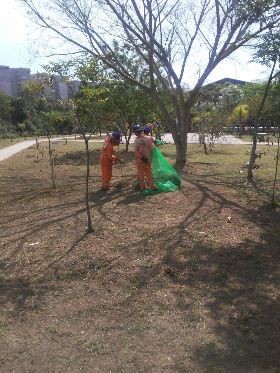 Três funcionários ao centro da imagem, rodeados por árvores, seguram uma rede com resíduos de grama.